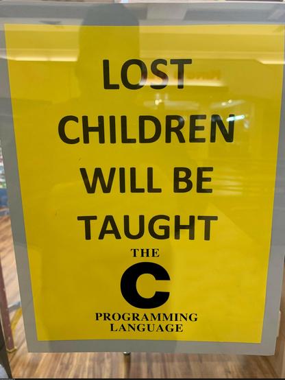 Big yellow sign on a glass door that says lost children will be taught the C programming language