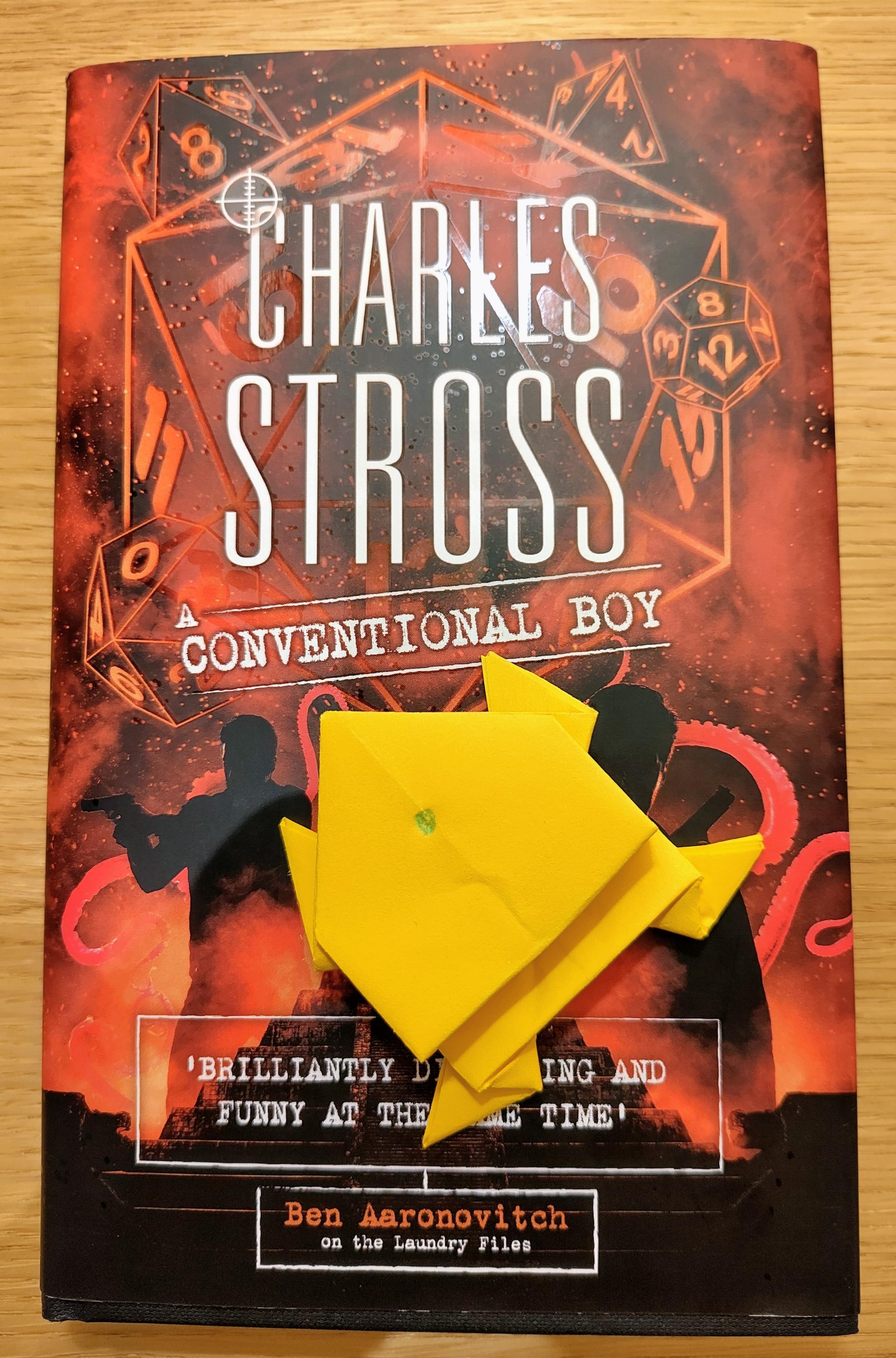 A gold origami frog with a green gem drawn between it's eyes on top of the book A Conventional Boy by Charles Stross